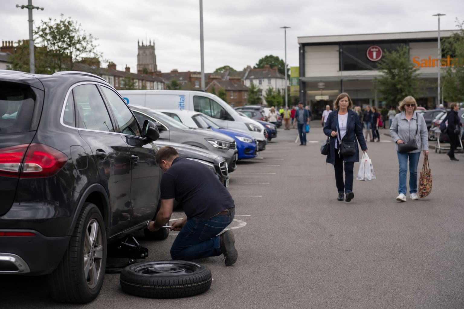 A man crouched at the back wheel of a car in a packed British town car park on an overcast Saturday, changing a flat tyre while shoppers walk past.