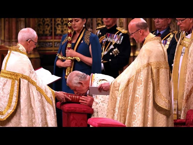 King Charles III bowed in prayer at his coronation in Westminster Abbey, hand resting on the Holy Bible, flanked by clergy in gold vestments with a ceremonial sword held upright behind him.